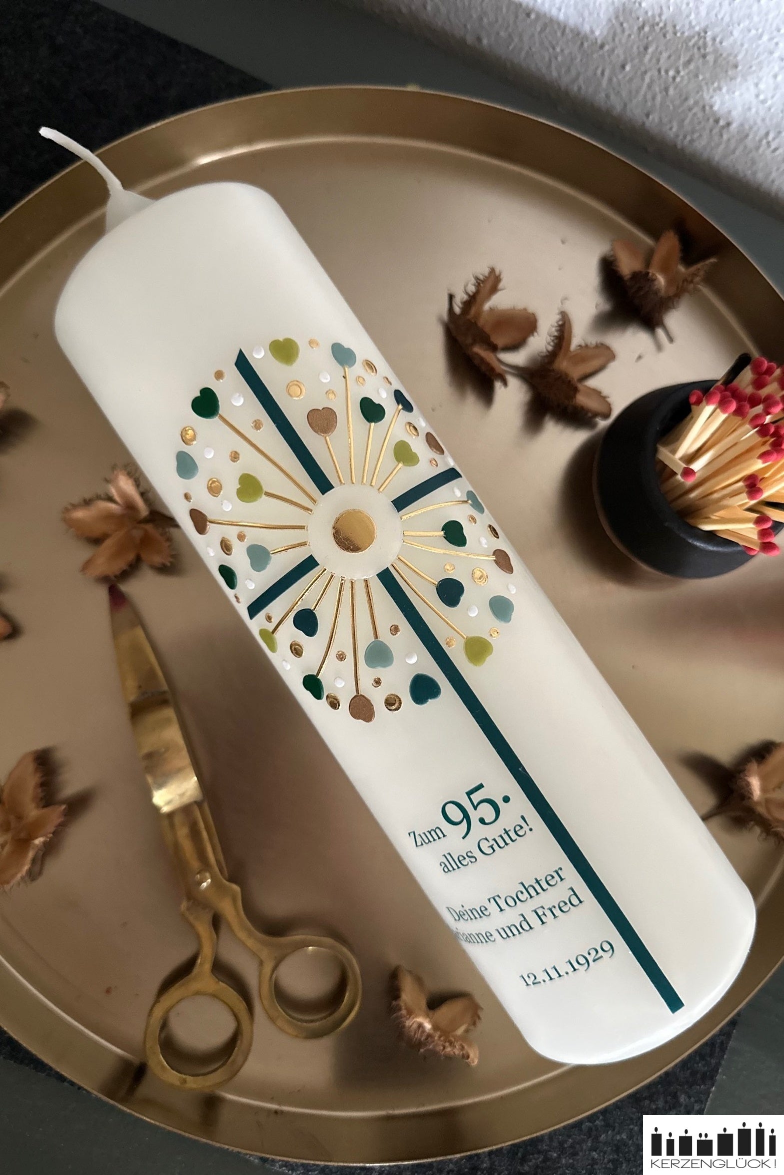 Geburtstagskerze Kreuz Pusteblume in Grüntönen und Goldfarben auf einem goldfarbenen Teller dekoriert mit herbstlichen Naturmaterialien, Streichhölzern und einer Messingschere zur Kerzenpflege