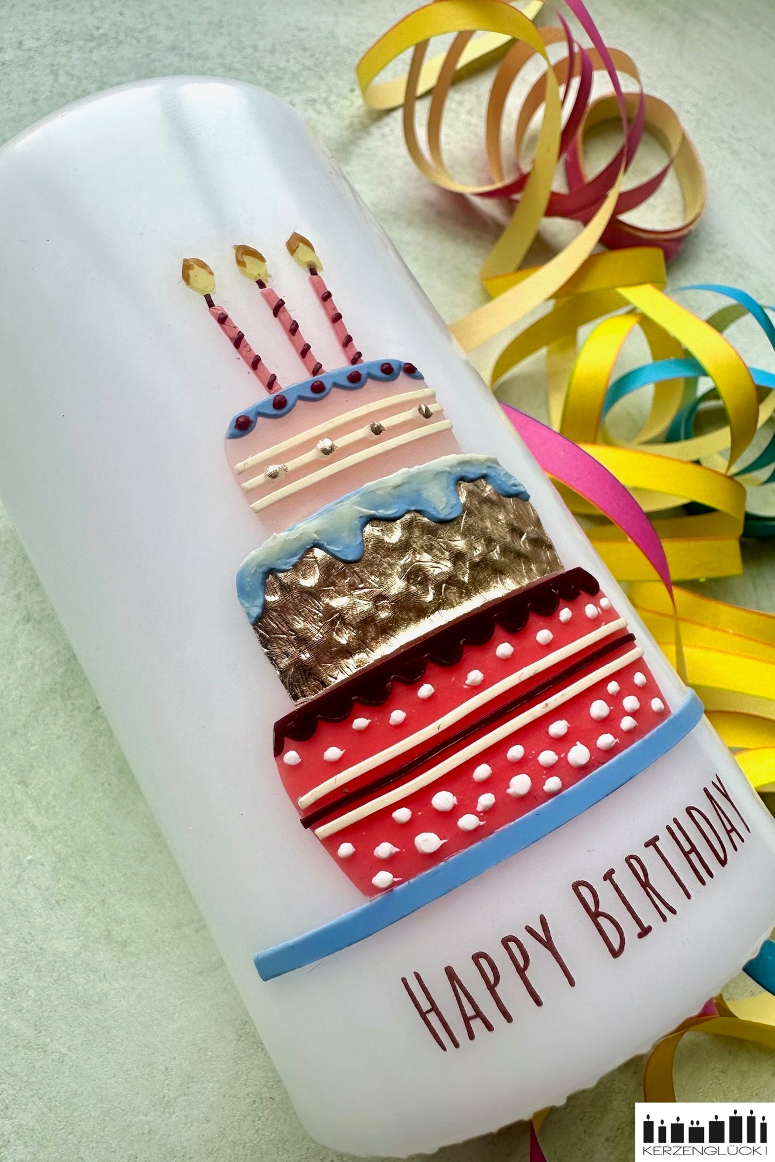 Eine Geburtstagskerze mit Torte und Kerzen verziert schräg liegend auf einer hellen Unterlage umrandet von Luftschlangen